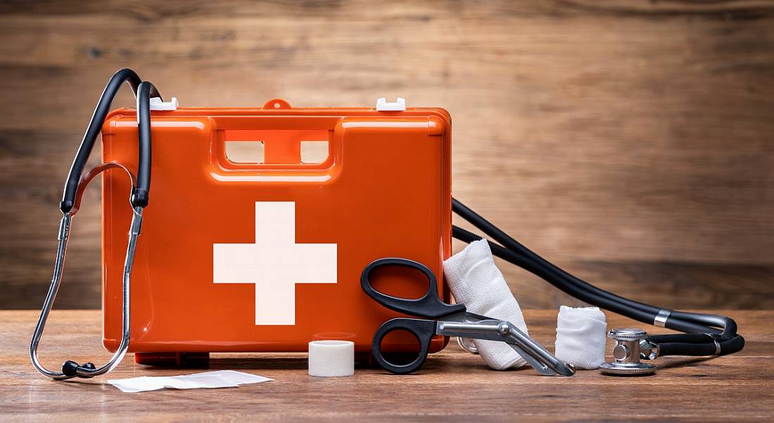 An orange first aid kit with a white plus sign sits on a wooden surface with some first aid objects scattered around, symbolising health and safety.