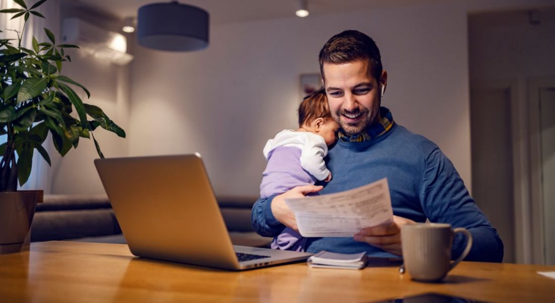 A man sits at a desk holding a baby in one arm and a sheet of paper in the other. There is a laptop on the desk in front of him.