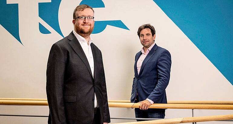 Two men standing separately from each other. One is holding on to a wooden handrail in the background.