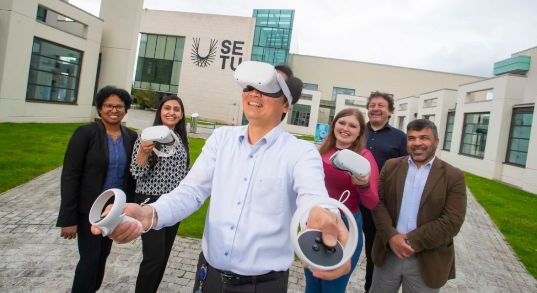 A group of people from SETU and DCU standing outside a building on SETU campus watching a man wearing a VR headset.
