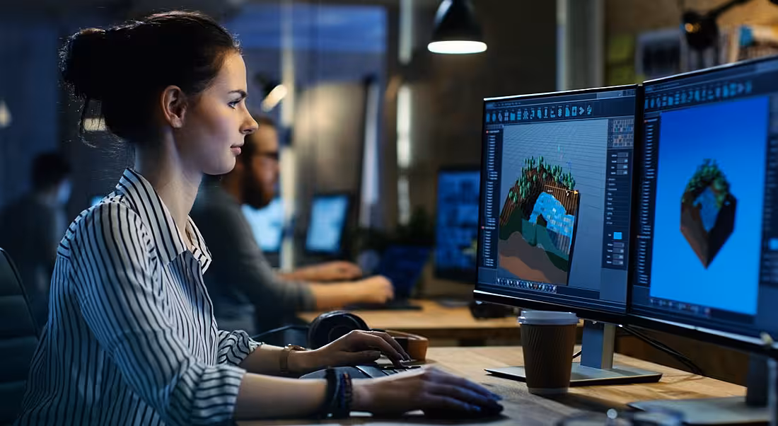 Developer working on her gaming development skills looking at a computer with two monitors in a dimly lit room.