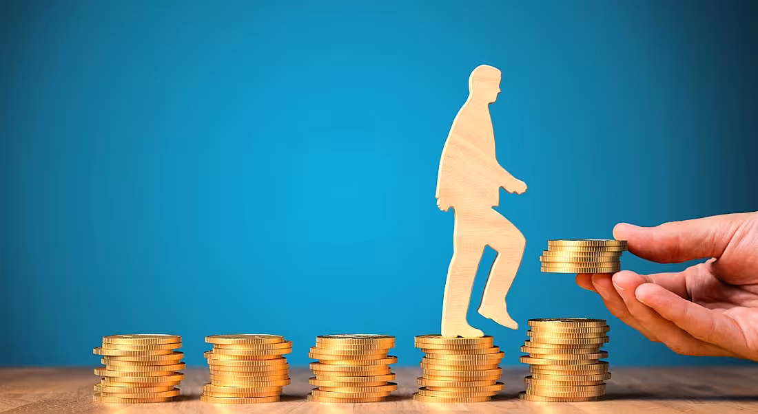 Piles of gold coins side by side gradually ascending with a wooden figure climbing on them and a hand adding more.