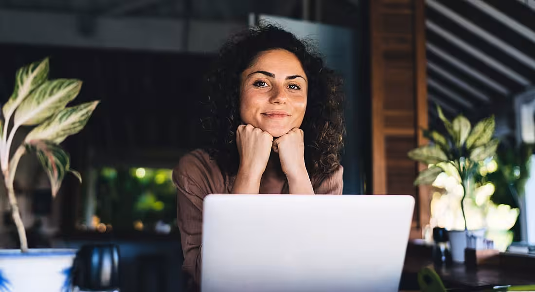 Woman who is sitting at a desk working remotely surrounded by her laptop and plants.