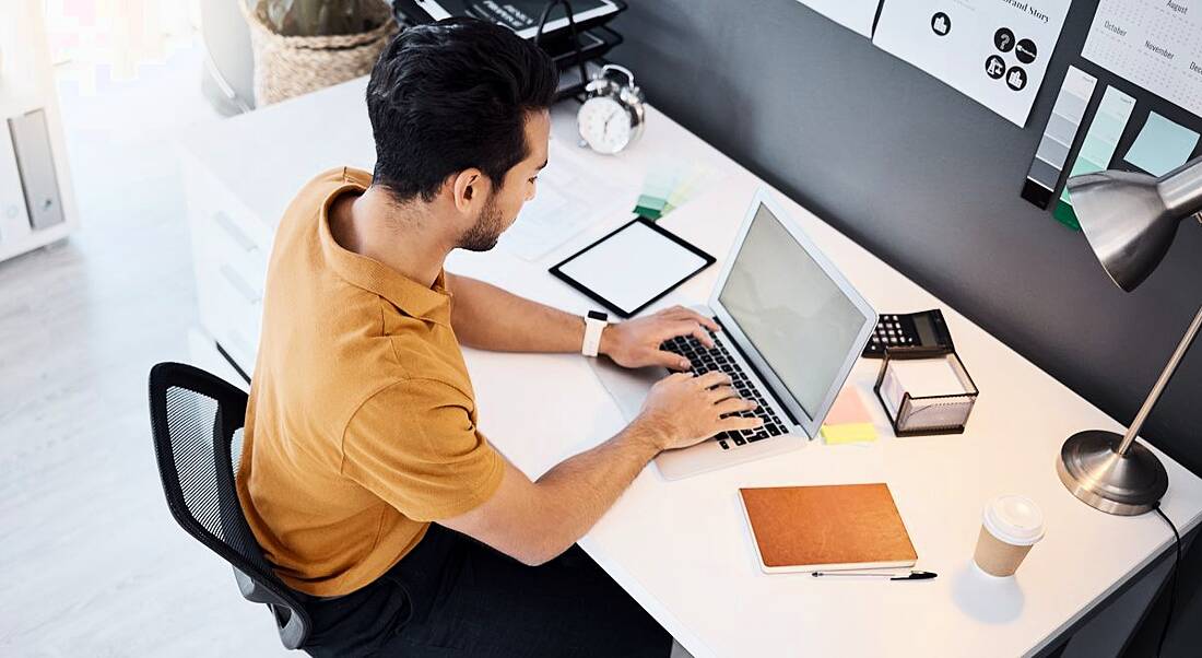 Man working on software on a laptop at a desk with notebooks and a lamp and a noticeboard in his surrounding area.