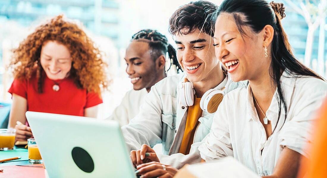 Young people sitting around a table together looking at a laptop and smiling like they are happy learning digital skills.