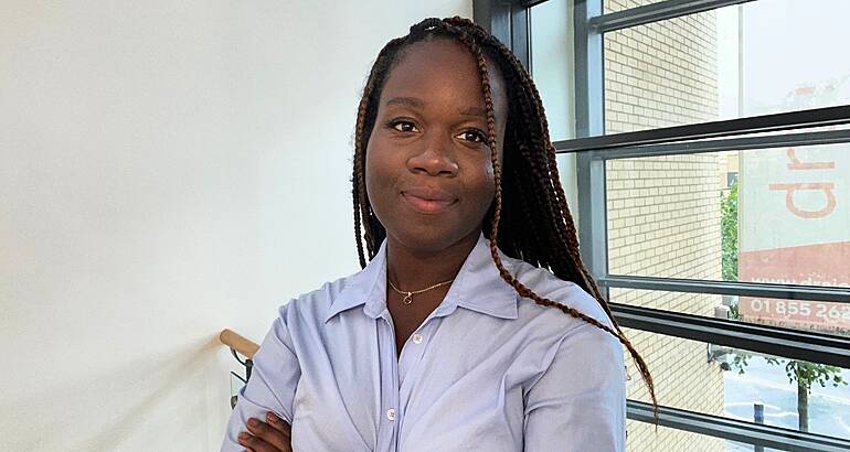 A woman wearing a light blue shirt smiles with her arms crossed in an office setting. She is Blessing Folayan, a radiographer and technology consulting analyst at Deloitte.