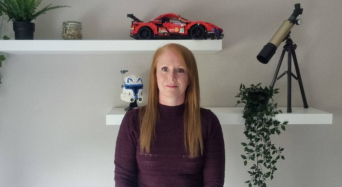 A woman wearing a maroon jumper stands smiling with a wall behind her. On the wall, there are two shelves with toys and plants on them. She is Lynn D'Arcy, a data centre engineer at Yahoo.