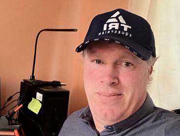 A man wearing a baseball cap smiles at the camera in a home office setting. There is a brown wall behind him as well as a computer. He is Shaun Haren, a software development intern at Fidelity Investments.