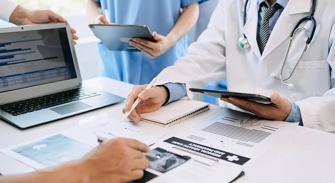 A group of medical staff stand around a desk full of medical records and documents, as well as a laptop.