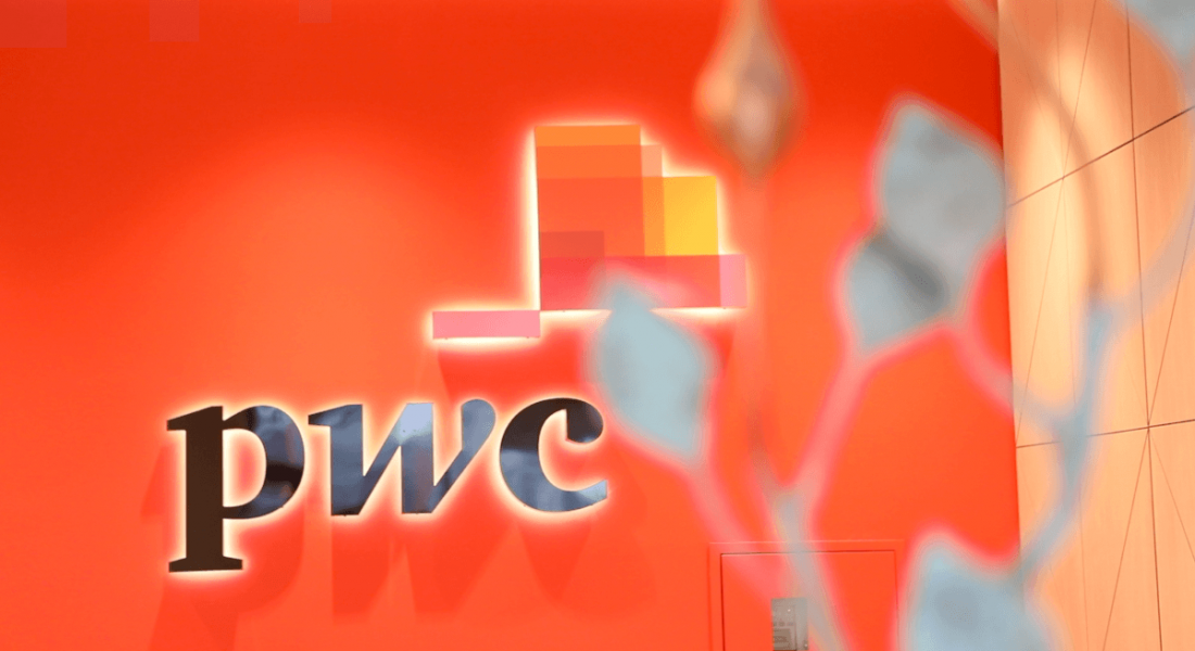 An image of the PwC logo on an orange wall. There is a plant in the foreground that is out of focus.