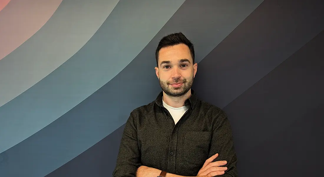 A man smiles while standing against a wall with his arms crossed. The wall is segmented into different shades of a blue gradient.