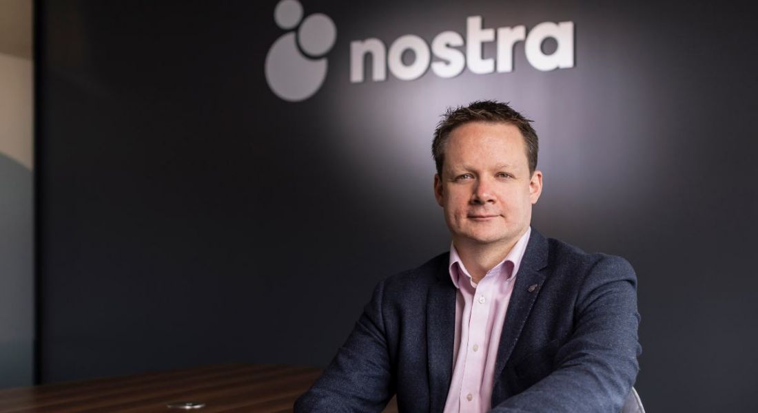 Nostra CEO Kevin O'Loughlin sitting in front of a grey wall that has Nostra branding on it.