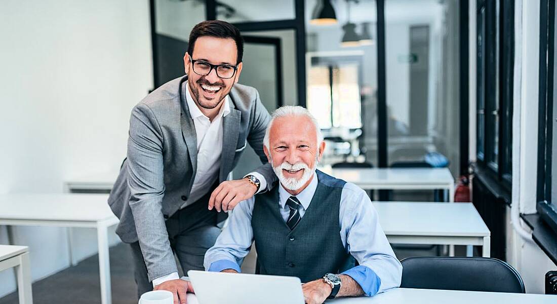 An older man sitting at a desk with a laptop while a younger man stands beside him.