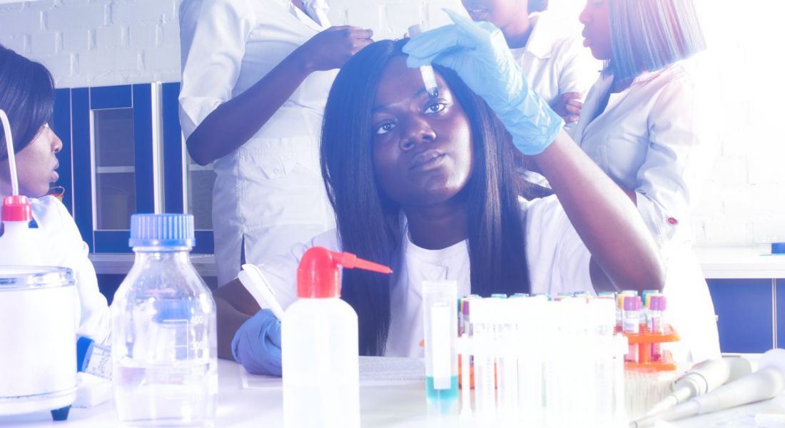 A young woman looking at a test tube in a lab environment with other biopharma workers standing behind her.
