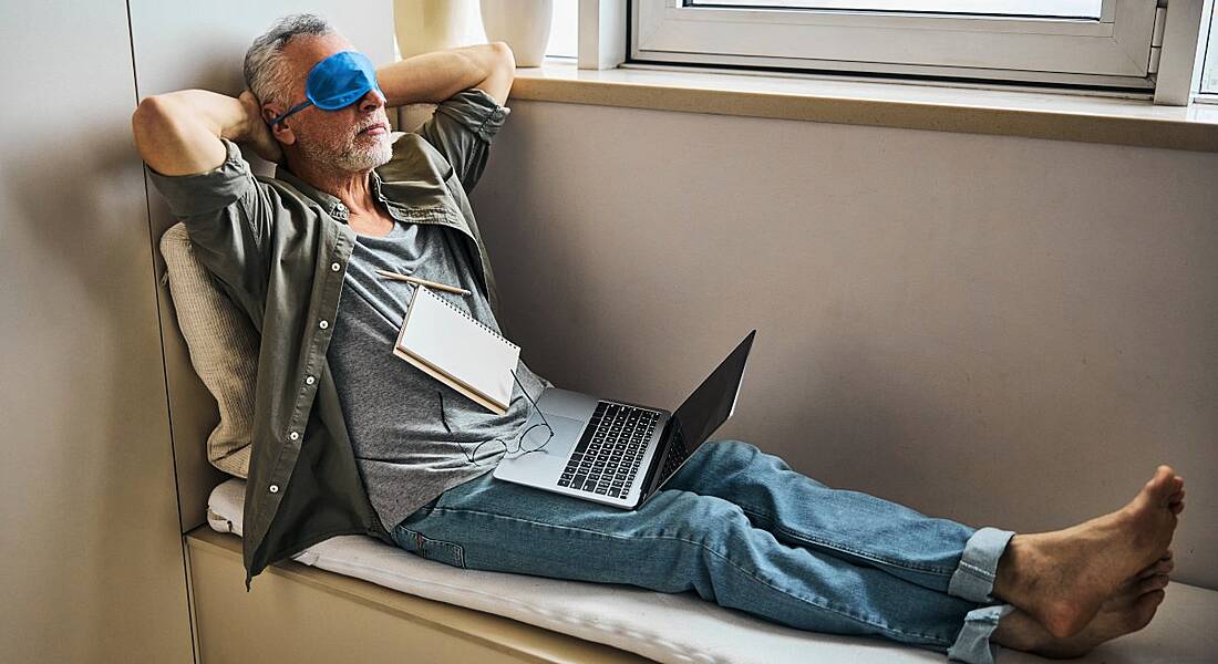 Man wearing an eye mask resting while sitting in a window seat in a relaxed pose with a laptop and notebook on his lap.