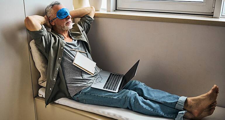 Man wearing an eye mask resting while sitting in a window seat in a relaxed pose with a laptop and notebook on his lap.