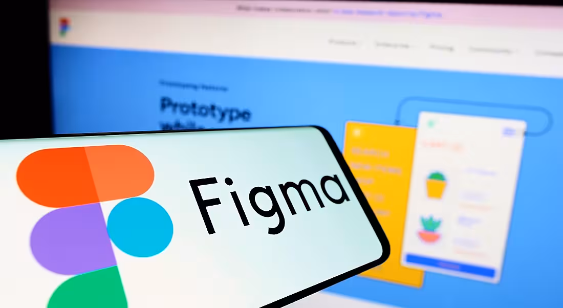 A phone with the Figma logo on it held up in front of a laptop with the Figma website.