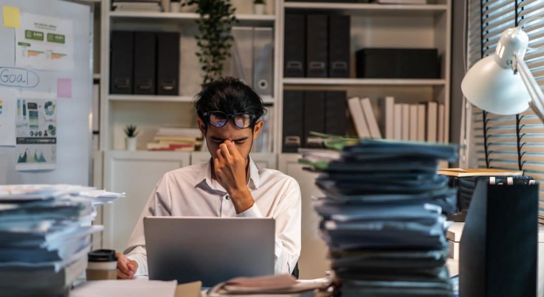 Worker sitting at his desk with his hand over his face feeling tired and surrounded by lots of paper documents and a computer.