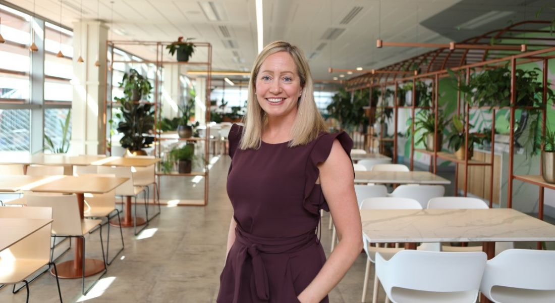 A woman wearing a maroon dress stands smiling in a brightly lit office setting with various tables and plants in the background. She is Aisling Curtis, market leader at PwC's strategic alliances team.
