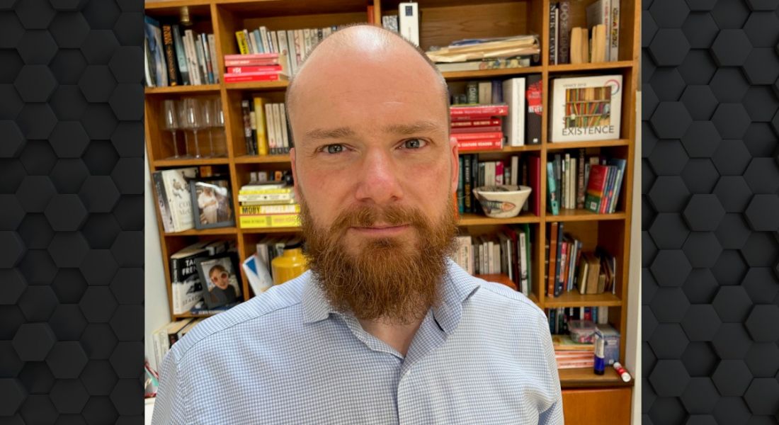 A bearded man wearing a light coloured shirt smiles at the camera in a home office setting. There are bookshelves behind him. He is Andrew Kealy, a software engineer at Fidelity Investments.