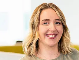 A woman smiles at the camera in front of a cream-coloured wall that has various framed posters hanging on it. She is Lisa Duncan, a human capital manager at Deloitte.