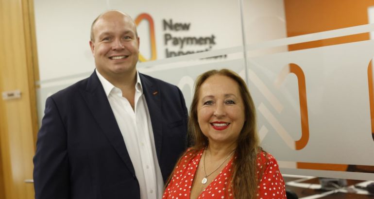 Carl and Nicole Churchill of New Payment Innovation standing against a white and orange wall with company branding on it.