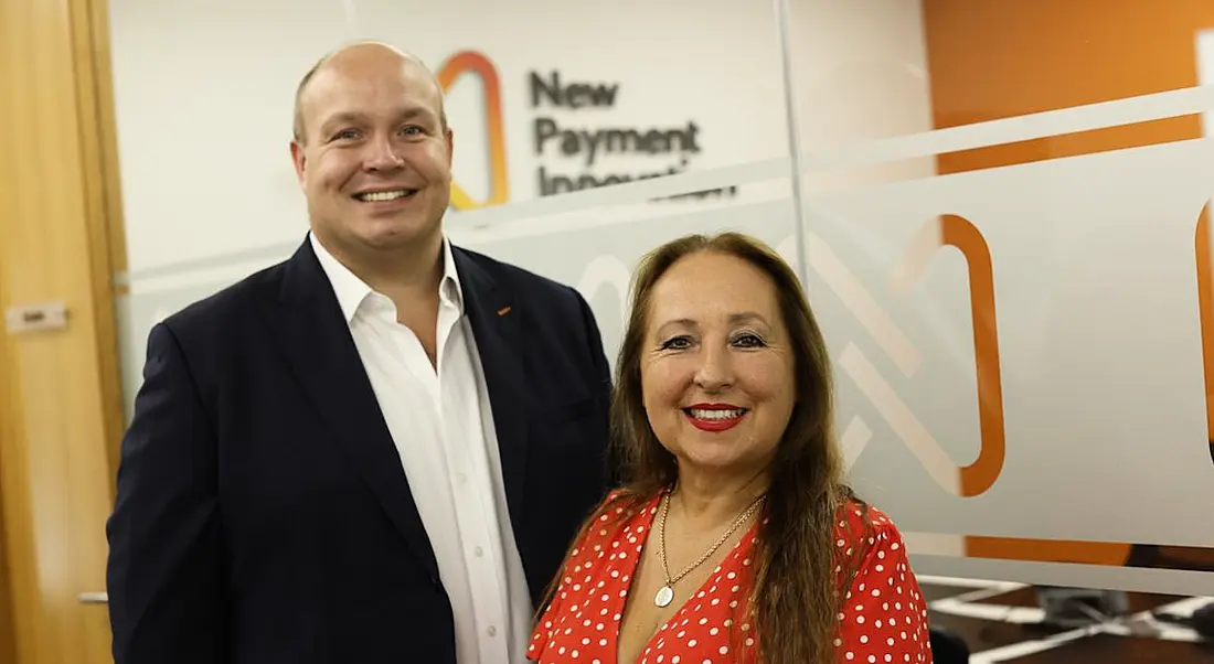 Carl and Nicole Churchill of New Payment Innovation standing against a white and orange wall with company branding on it.