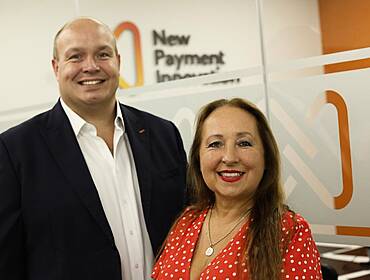 Carl and Nicole Churchill of New Payment Innovation standing against a white and orange wall with company branding on it.