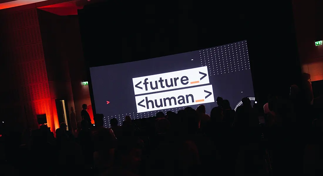 A darkened room with a large seated crowd in front of a large screen with the Future Human logo on display.