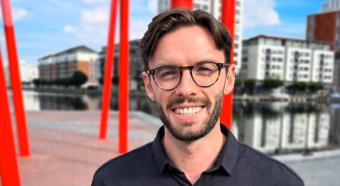 A man wearing a black shirt and glasses smiles at the camera in an outside urban setting. He is Geoff Allen, a management consulting analyst at Accenture.