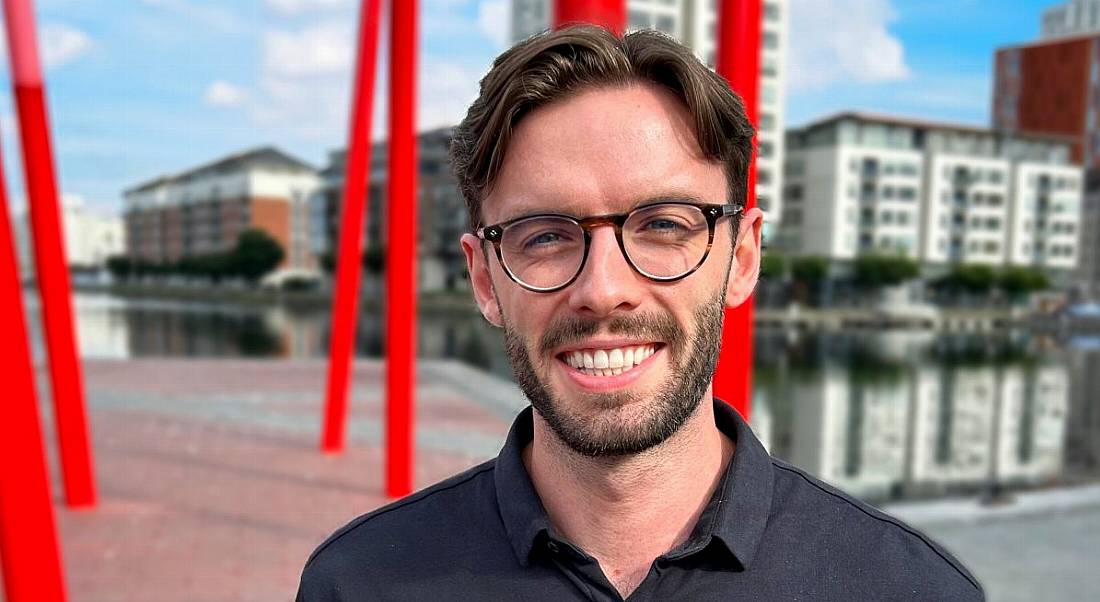 A man wearing a black shirt and glasses smiles at the camera in an outside urban setting. He is Geoff Allen, a management consulting analyst at Accenture.