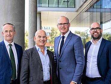 Four men stand in a line outside a modern building on a sunny day smiling at the camera. It is the new Integrity360 building.