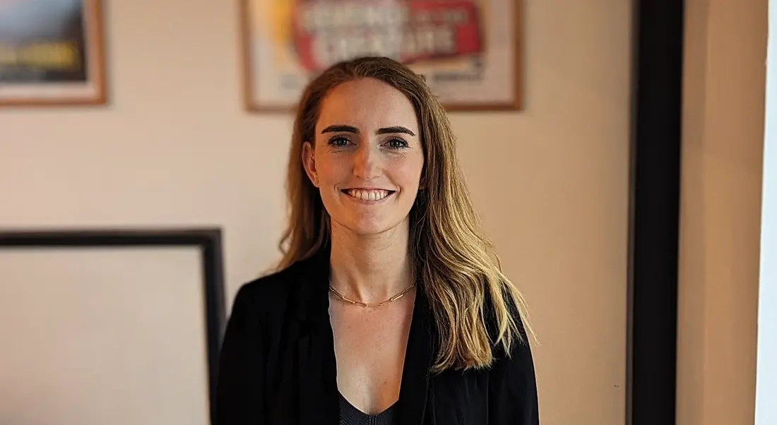 A woman smiles at the camera in front of a cream-coloured wall that has various framed posters hanging on it. She is Lisa Duncan, a human capital manager at Deloitte.