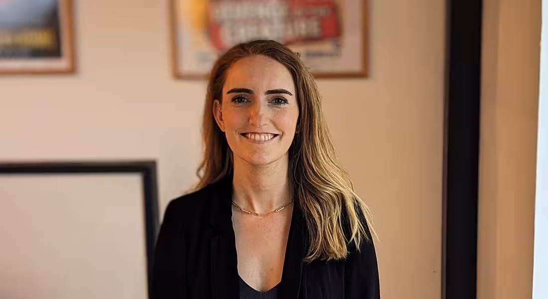 A woman smiles at the camera in front of a cream-coloured wall that has various framed posters hanging on it. She is Lisa Duncan, a human capital manager at Deloitte.