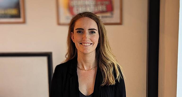 A woman smiles at the camera in front of a cream-coloured wall that has various framed posters hanging on it. She is Lisa Duncan, a human capital manager at Deloitte.