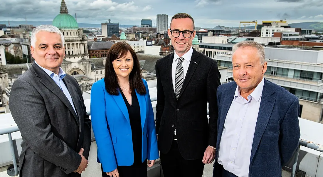 Four people standing on a balcony on the rooftop of a building with a city and buildings and a skyline visible behind them.