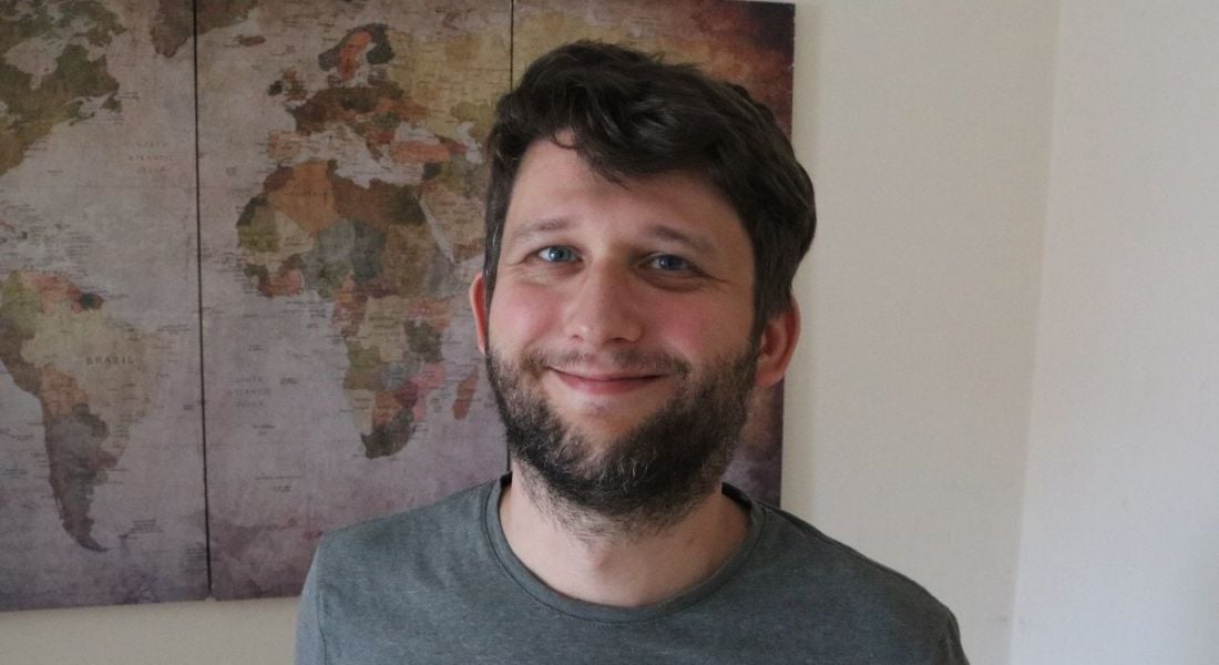 A man wearing a dark grey t-shirt smiles at the camera in front of a map of the world hanging on a white wall. He is Robert Soldan, a software engineer at Yahoo.