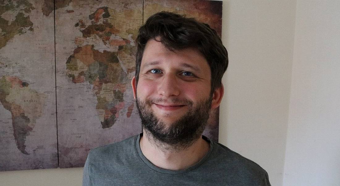 A man wearing a dark grey t-shirt smiles at the camera in front of a map of the world hanging on a white wall. He is Robert Soldan, a software engineer at Yahoo.