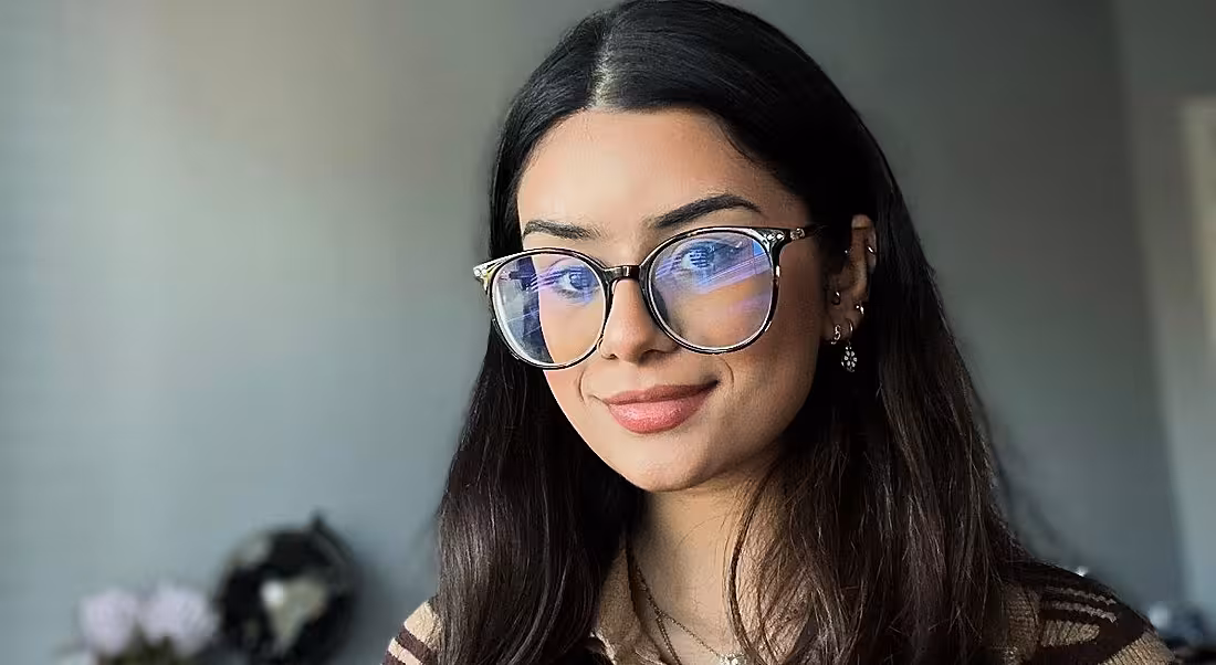 A woman with long dark hair wearing glasses smiles at the camera in front of a grey wall. She is Saira Mehdi, a consultant at Grant Thornton.