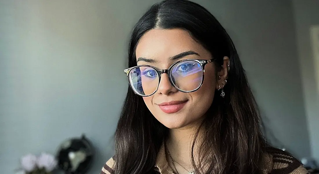 A woman with long dark hair wearing glasses smiles at the camera in front of a grey wall. She is Saira Mehdi, a consultant at Grant Thornton.