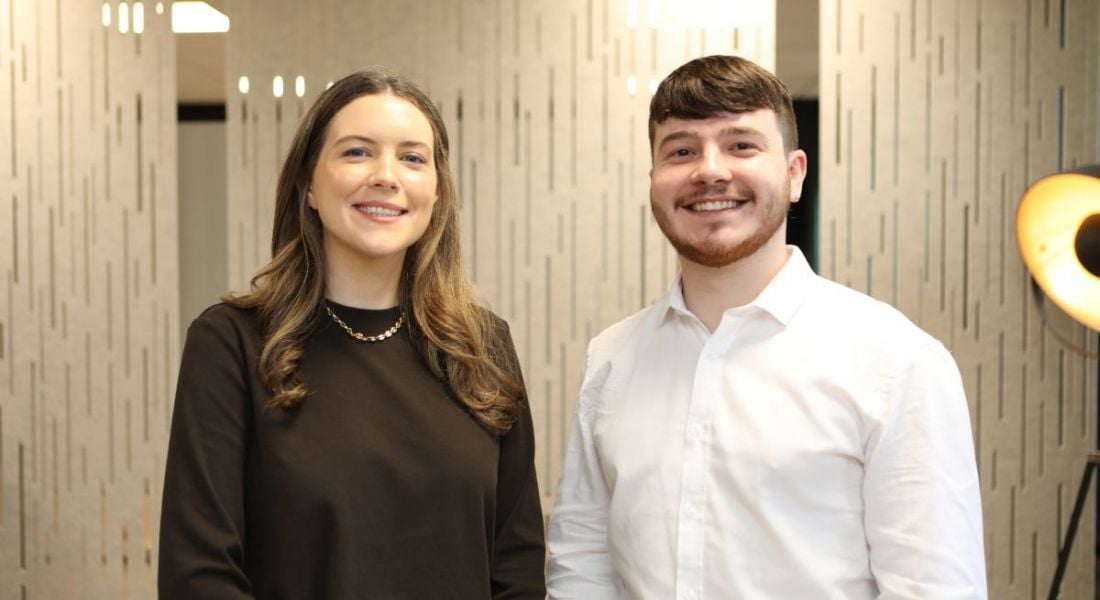 A woman and a man stand next to each other smiling at the camera in an office setting. They are Sinéad Coughlan and Robyn Duggan.