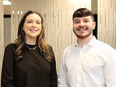 A woman and a man stand next to each other smiling at the camera in an office setting. They are Sinéad Coughlan and Robyn Duggan.