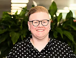 A woman smiles at the camera in front of a cream-coloured wall that has various framed posters hanging on it. She is Lisa Duncan, a human capital manager at Deloitte.