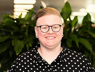 Sarah Comiskey, senior technology consultant at BearingPoint, stands smiling at the camera with a large plant blurred behind her.