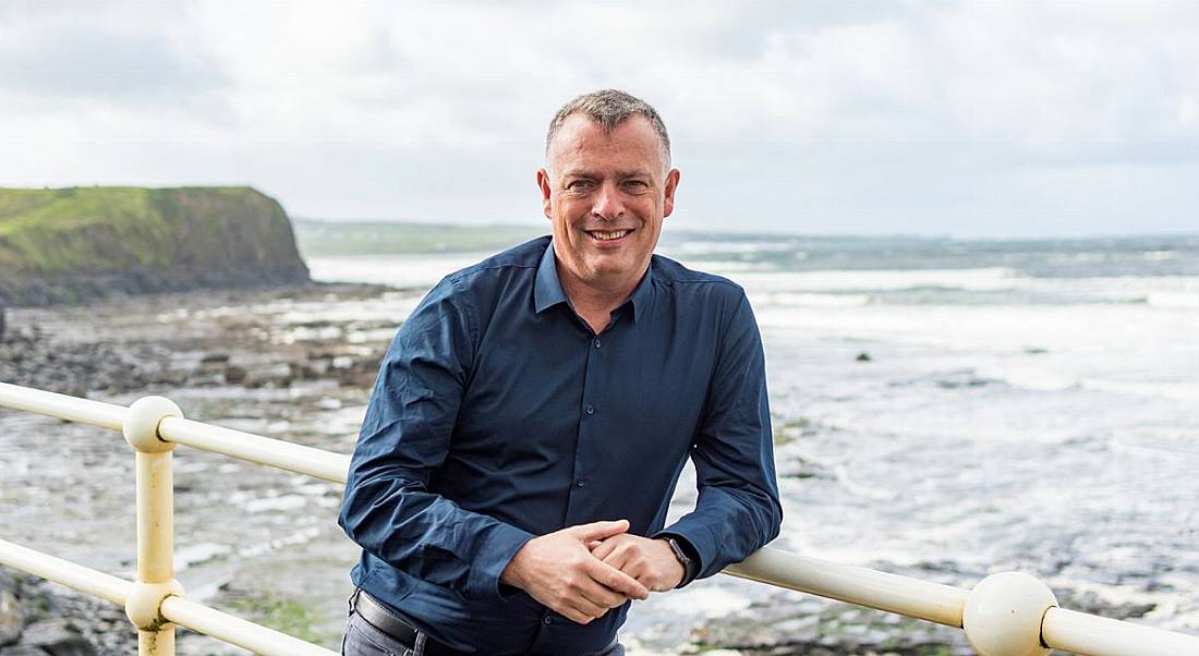 A man called Adam Coleman leans on a railing overlooking a rocky beach with a cliff and waves featuring in the background.