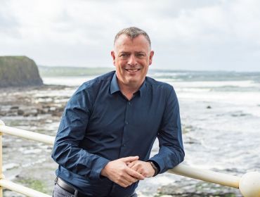 A man called Adam Coleman leans on a railing overlooking a rocky beach with a cliff and waves featuring in the background.