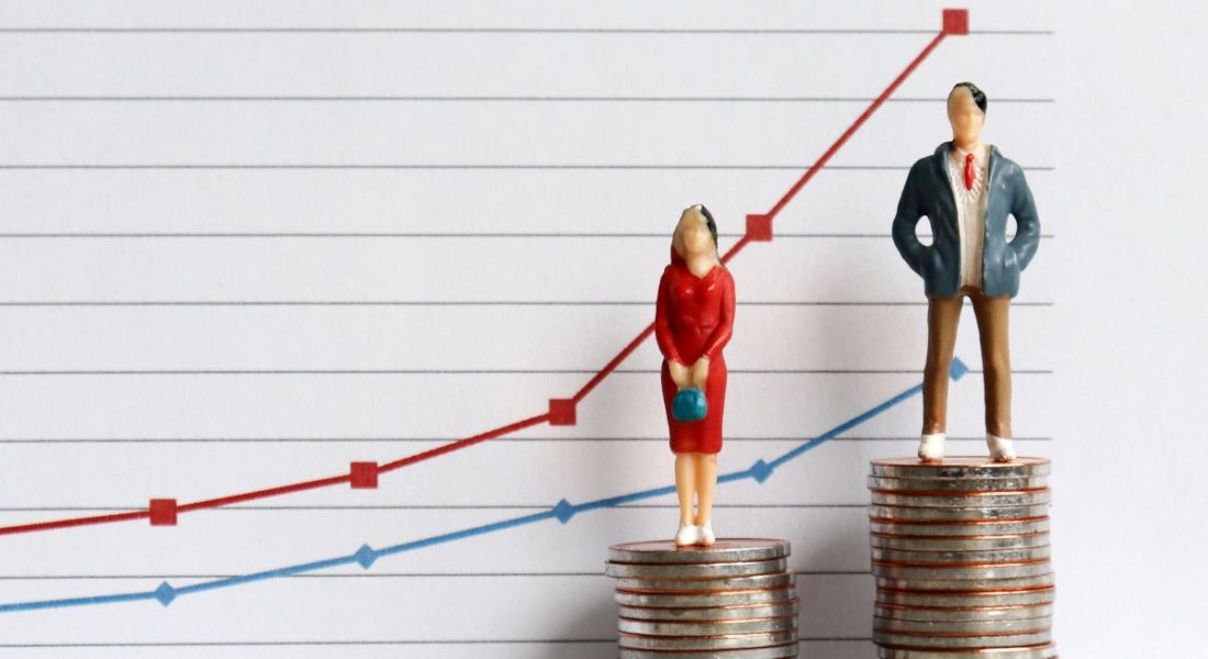 Figurines of a man and woman standing on stacks of coins with a graph behind them, representing gender pay gap concepts.