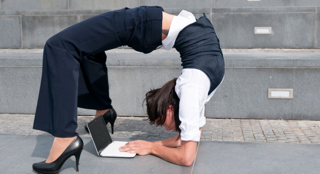 Woman wearing office attire contorts her body so that she is literally bending over backwards to work on a laptop.