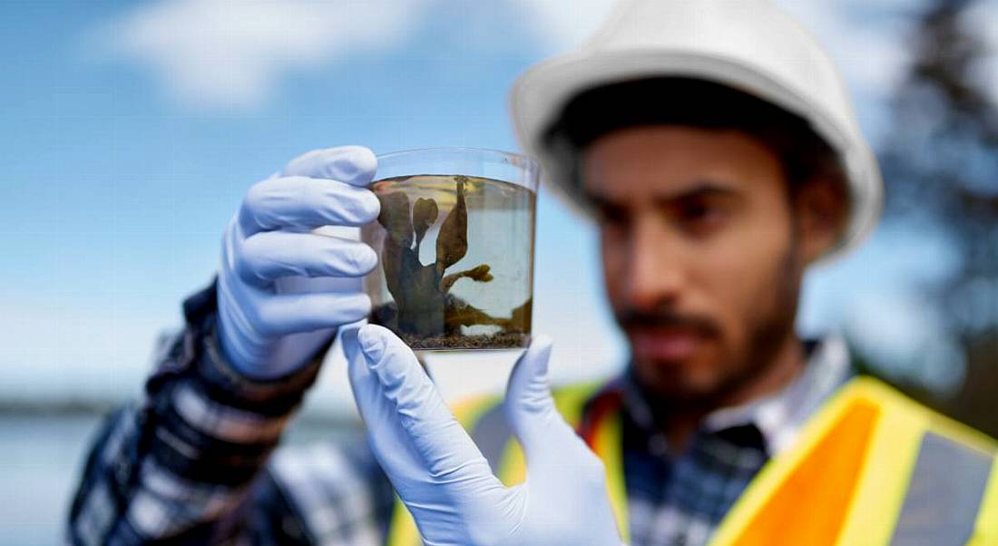 Marine scientist holding up a seaweed specimen in a jar examining it. He is wearing gloves and protective gear.