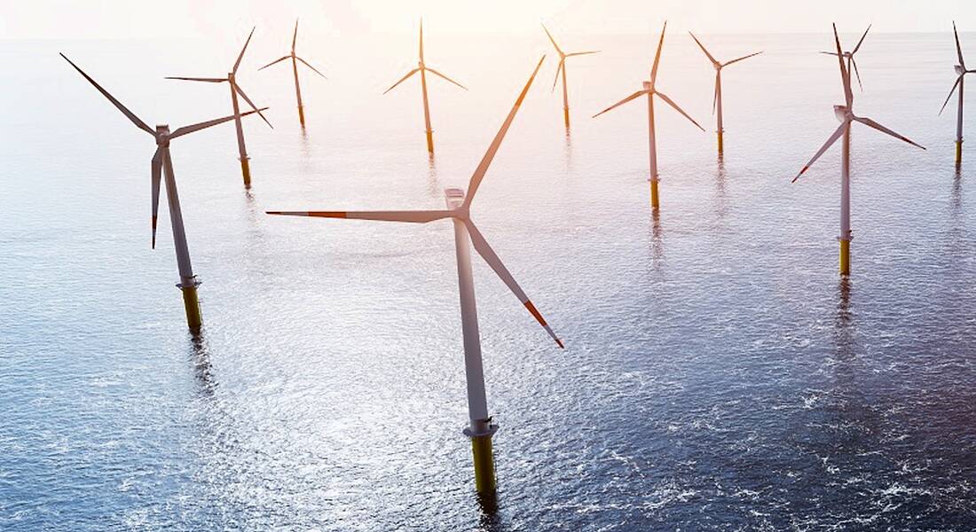 An offshore windfarm, with multiple turbines in the sea, with the sun shining in the distance.